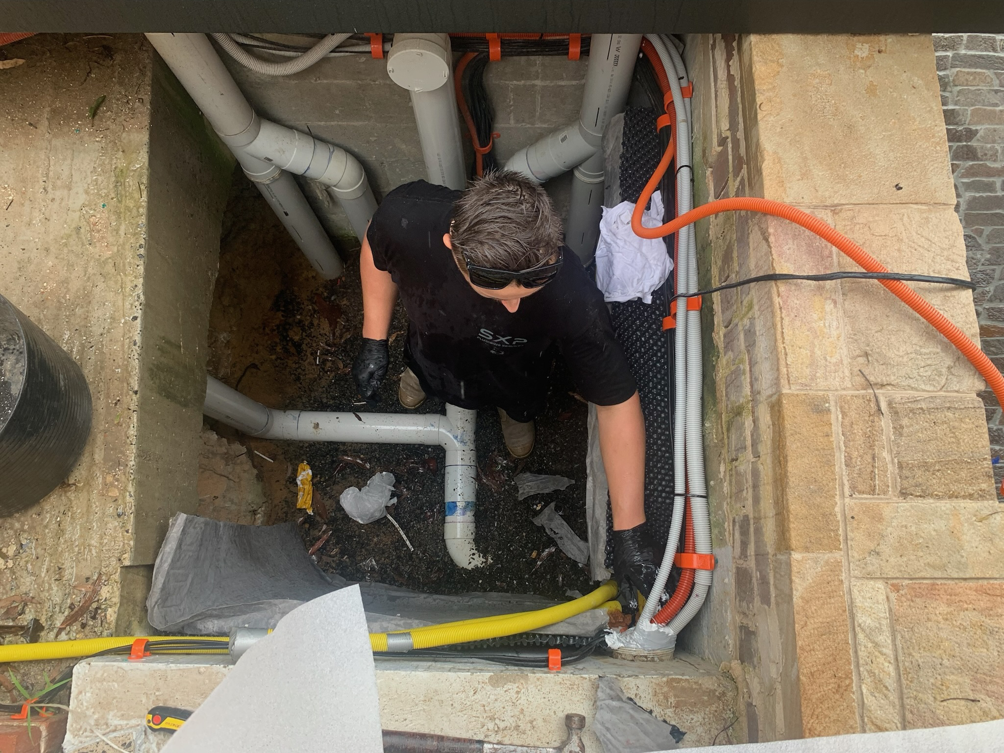A person in black clothing and gloves works with pipes and cables in a confined outdoor utility area surrounded by stone and concrete walls, expertly handling tasks typical for Strata Plumbing professionals.
