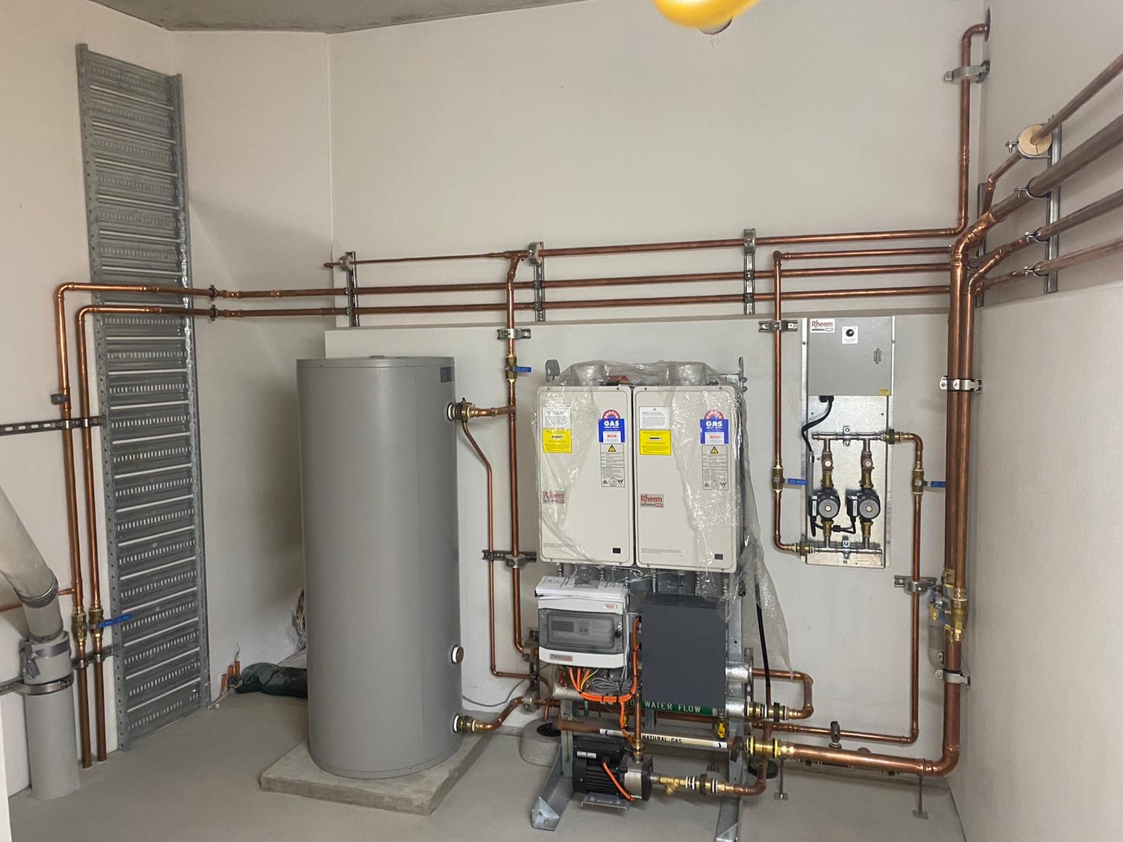 A utility room with a water heater tank, multiple wall-mounted boilers, exposed copper piping, a control panel, and various valves and gauges—ideal for troubleshooting hot water issues efficiently.
