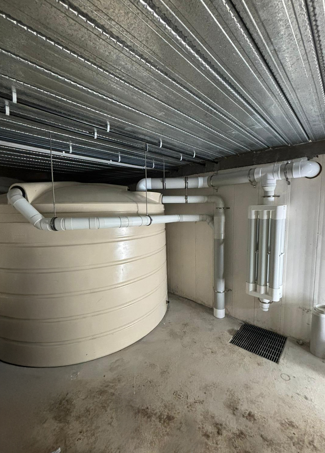 Beige water storage tank with white PVC pipes and filters for stormwater, installed indoors under corrugated metal ceiling.