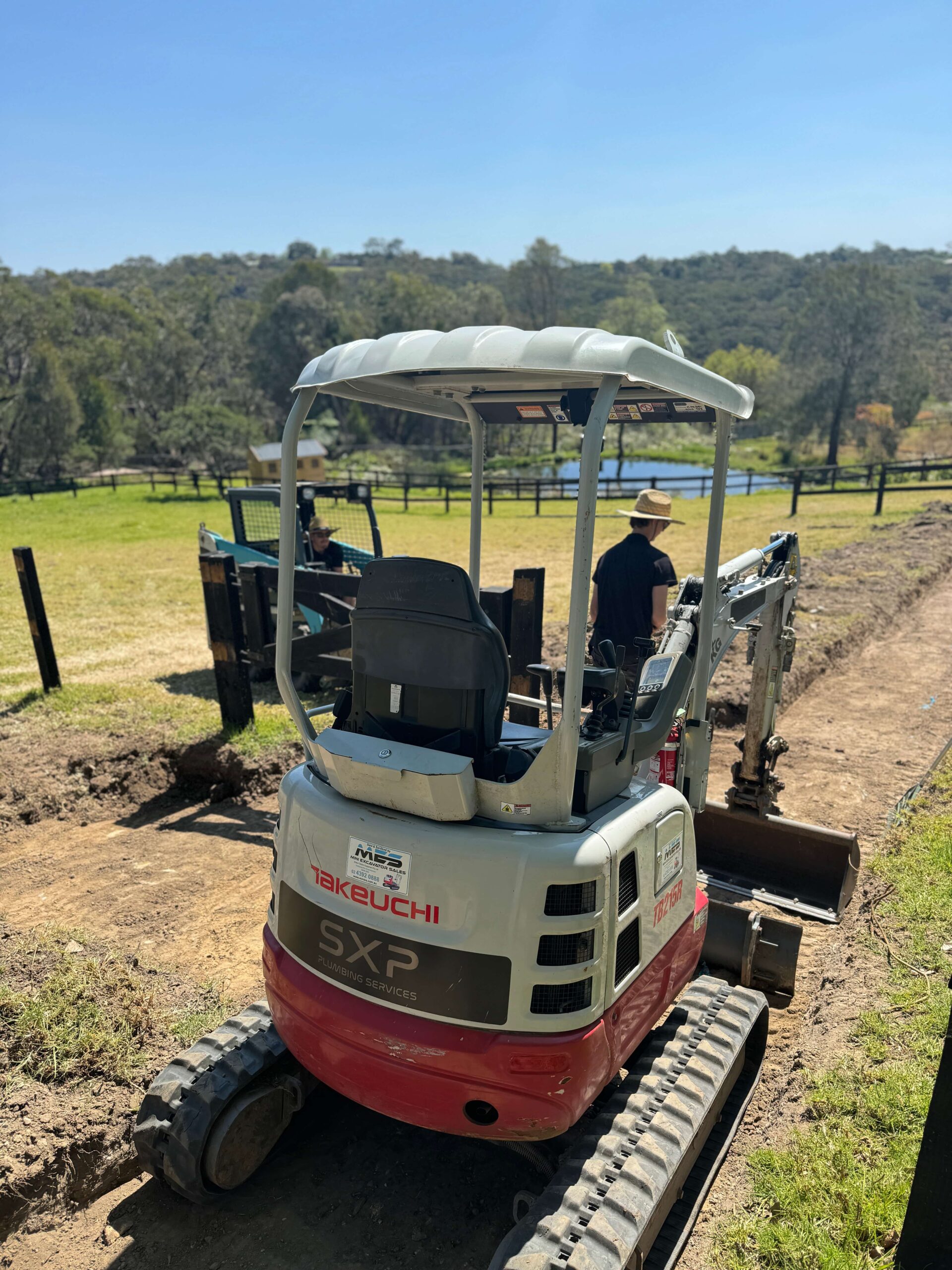 Integrated plumbing expert uses a small Takeuchi excavator on a dirt path as another person watches, both wearing hats.