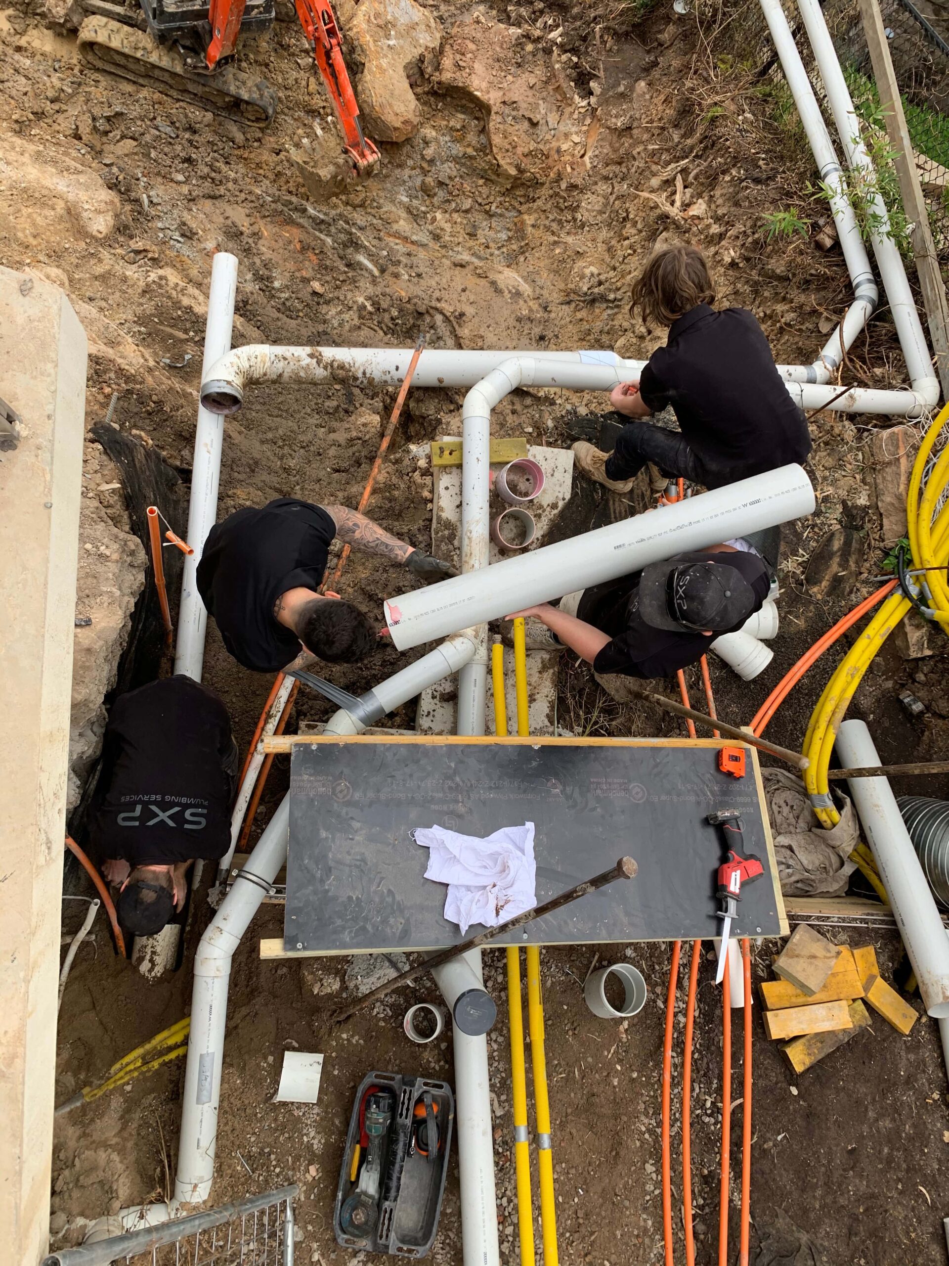 four workers connect white PVC pipes in a trench for drainage solutions, with tools, cables and hoses at a construction site.