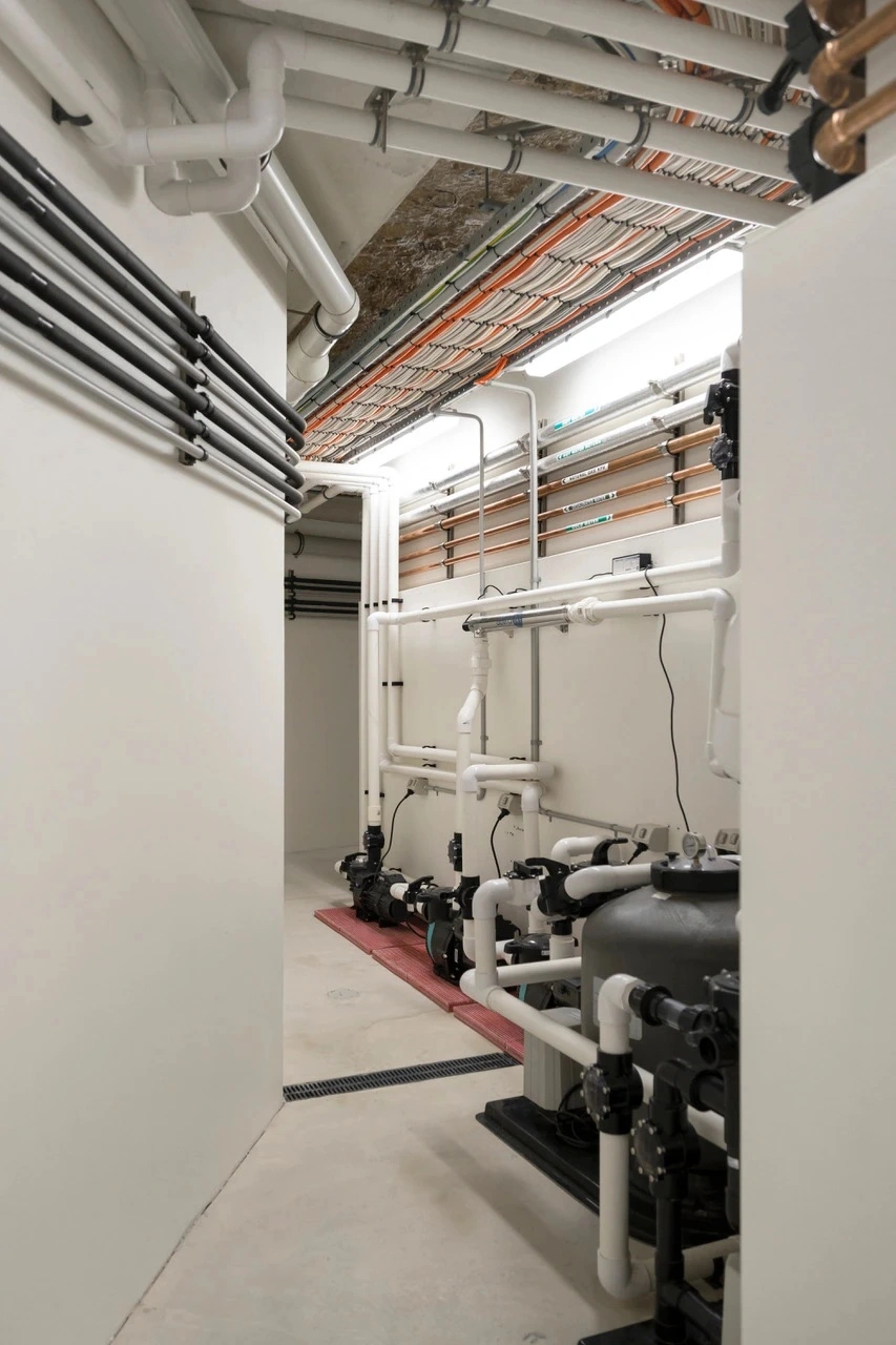 geotagged-02-Commercial - SXP Plumbing - SXP Plumbing Mechanical room with white PVC pipes, valves, and multiple wall-mounted pumps along the side wall.